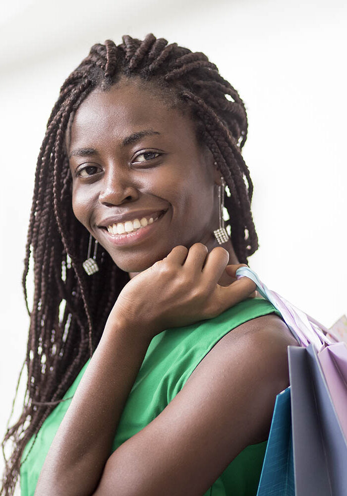 Portrait of happy young African American woman wearing braids holding shopping bags, looking at camera and smiling indoors
